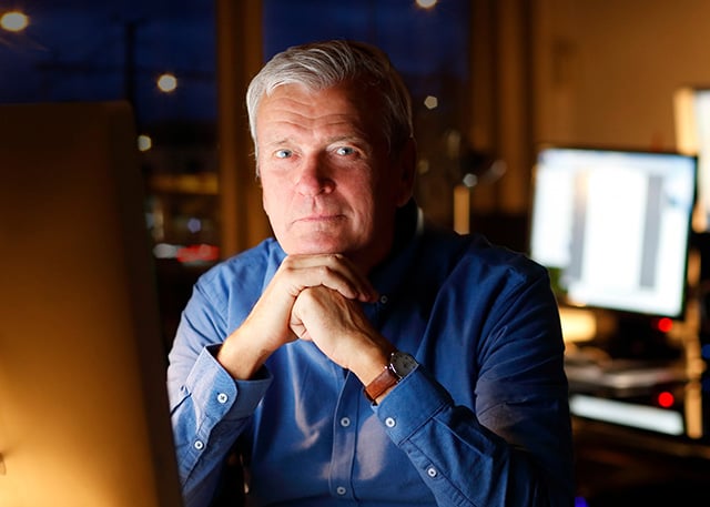 code-sign-4 A confident older man in a blue shirt sitting at a desk in a dimly lit office, with computer screens in the background, symbolizing experience in cybersecurity or IT leadership.