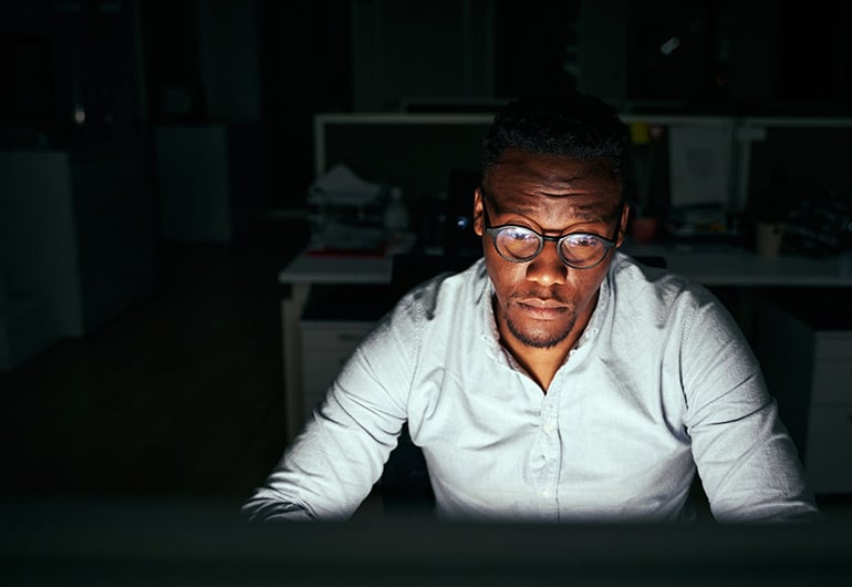 CS-hero A focused man wearing glasses and a light-colored shirt working late at night in a dimly lit office, illuminated by his computer screen, symbolizing cybersecurity, coding, or IT operations