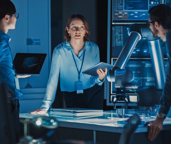 Diverse Team of Engineers with Laptop and a Tablet Analyse and Discuss How a Futuristic Robotic Arm Works and Moves a Metal Object. They are in a High Tech Research Laboratory with Modern Equipment. Manufacturing